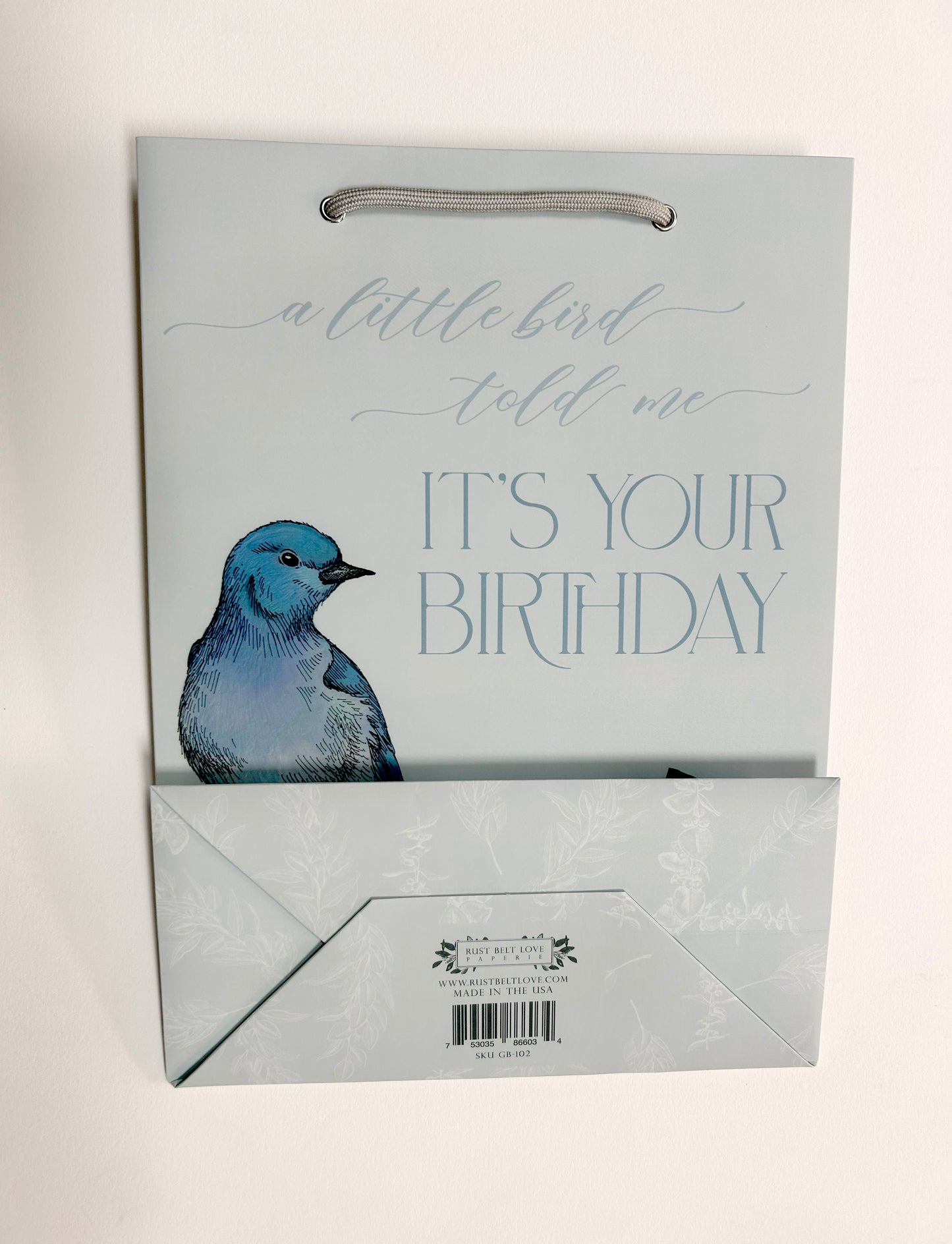 Picture of a blue birthday gift bag with florals and a bird that reads "A little bird told me it's your birthday" by Rust Belt Love, layed flat with the bottom of the bag visible as well.