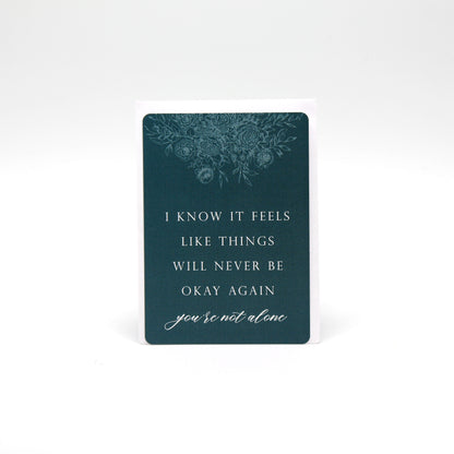 Dark teal floral minicard that reads "I know it feels like things will never be okay again you're not alone" by Rust Belt Love