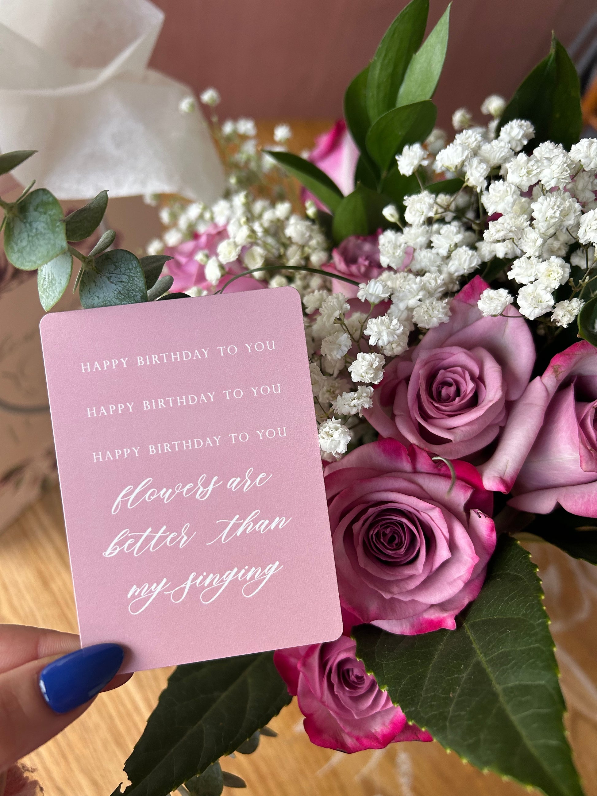 Style shot of pink minicard that reads "Happy birthday to you, Happy birthday to you, Happy. birthday toyou, flowers are better than my singing", in a bouquet of flowers, by Rust Belt Love.