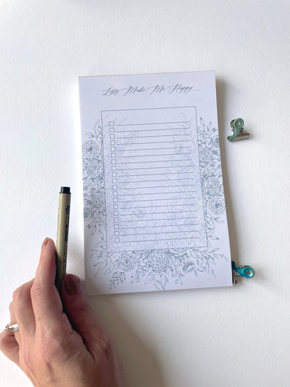 Style shot of 5.5 by 8.5 notepad with lined sheets with blue florals that says "Lists make me happy" by Rust Belt Love